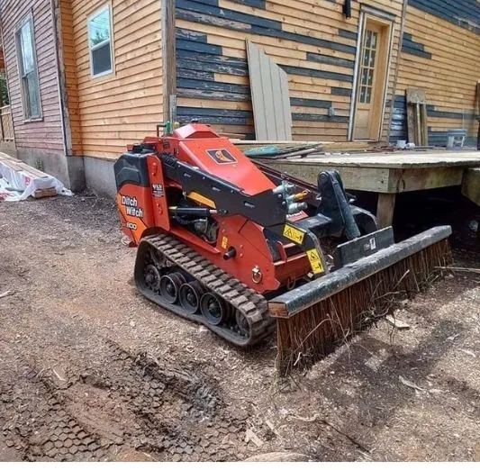 Compact track loader with a brush attachment clearing the ground beside a house