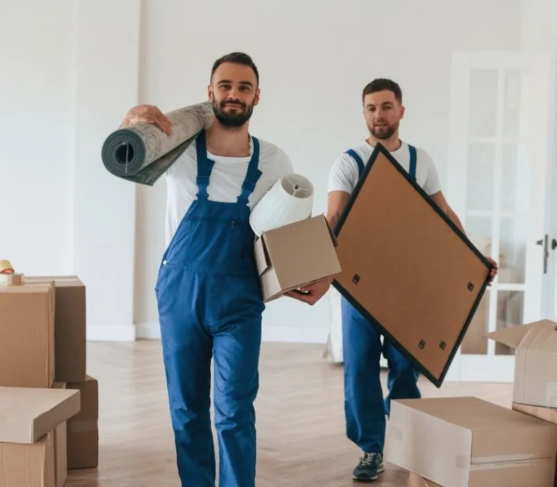 Two movers carrying a rolled rug, a frame and boxes