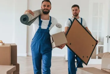 Two movers in blue overalls carrying a rug, boxes and a picture frame