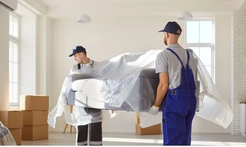 Movers carrying a wrapped sofa out of a bright room