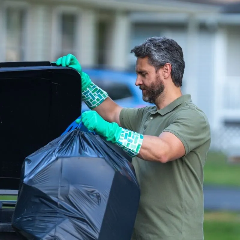 Man in green rubber gloves putting a full trash bag into a garbage bin