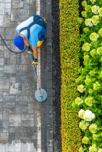 Overhead view of a worker pressure washing a paver walkway beside a garden